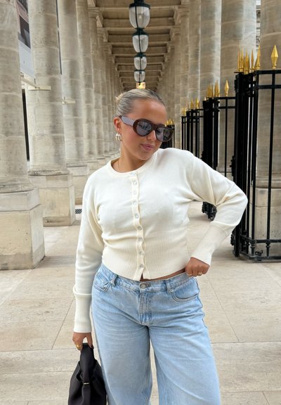 Joven mujer con gafas de sol lleva un suéter blanco abotonado y vaqueros azul claro, de pie junto a columnas de piedra y una valla negra con puntas doradas.