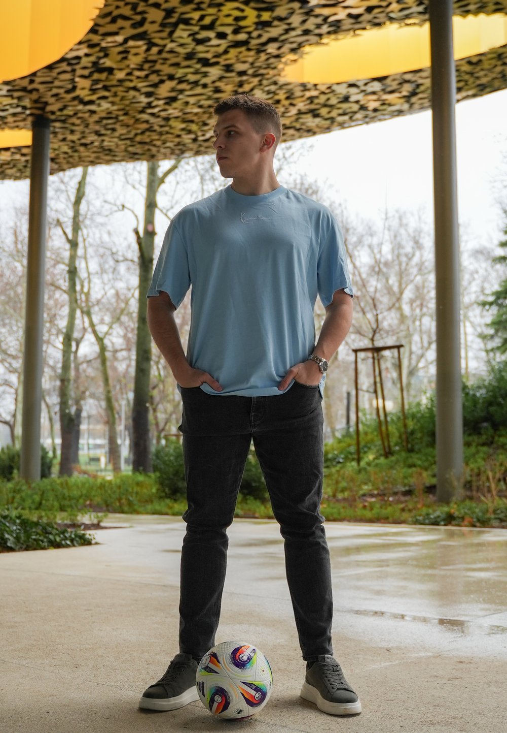 Ljusblå t-shirt med kort ärm, mörka denimjeans, grå sneakers; står på en slät yta med en flerfärgad fotboll vid fötterna.