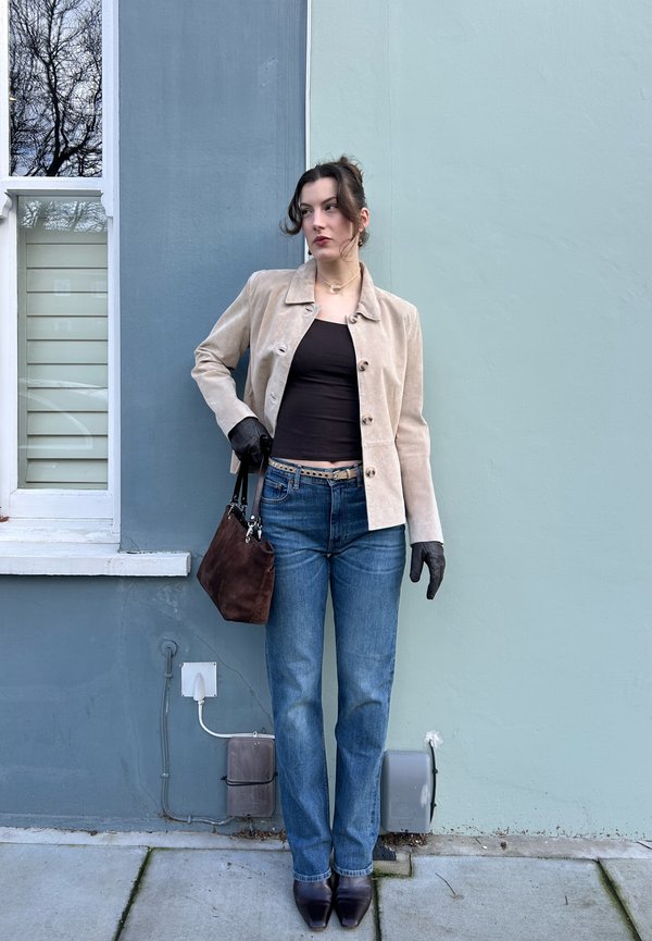 Donna con giacca beige, top nero, jeans blu, guanti neri e stivali, che tiene una borsa marrone, in piedi contro un muro blu-grigio vicino a una finestra.