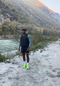 Idrettsbekledning: svart vest, grønn langermet topp, svarte shorts, hvite sokker, lyse gule joggesko. Naturlandskap med vann og trær.