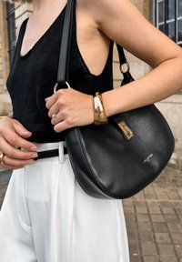 Schwarze Lederhandtasche mit einer gebogenen Form, ausgestattet mit einem goldenen Reißverschluss und Hardware-Akzenten, getragen über einer weißen hoch taillierten Hose.
