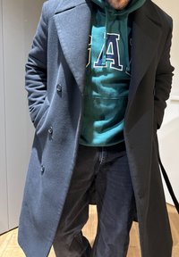 Άνδρας που φοράει σκούρο γκρι μακρύ παλτό και πράσινη φούτερ με μεγάλες γράμματα "UA", με  τα χέρια στις τσέπες του παλτού, στέκεται σε ξύλινο πάτωμα.