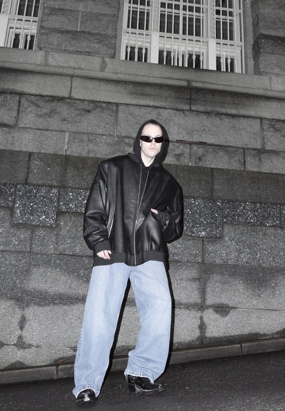 Homme portant une veste en cuir noire surdimensionnée, un jean large bleu clair, des bottes noires et des lunettes de soleil, debout dans la rue contre un mur en pierre la nuit.