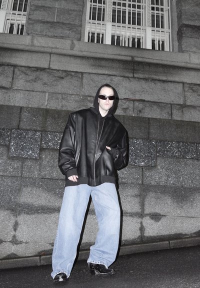 Hombre con chaqueta de cuero negra extragrande, vaqueros anchos de color azul claro, botas negras y gafas de sol, de pie en la calle frente a una pared de piedra por la noche.