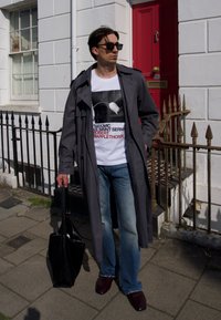 Grå trenchcoat over en hvit grafisk t-skjorte, blå flarejeans og glansfulle mørke sko. Holder en svart teksturert totebag. Rød dør i bakgrunnen.