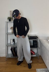 Maglietta nera a maniche lunghe, pantaloni tuta grigi e calzature nere. Ambientazione minimalista con una pianta, uno scaffale e un giradischi.