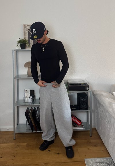 Camisa negra de manga larga, pantalones de chándal grises y calzado negro. Ambiente minimalista con una planta, una estantería y un tocadiscos.