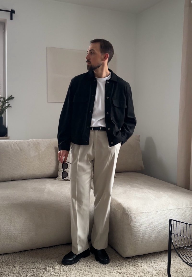 Uomo in piedi al chiuso che indossa una giacca nera, camicia bianca, pantaloni beige, tiene in mano degli occhiali da sole e guarda di lato vicino a un divano beige.