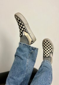 Black and white checkerboard slip-on shoes showcased with grey socks, resting on denim-clad legs against a neutral backdrop.