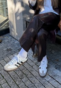 Oseba sedi na klopi, oblečena v bele nogavice, bele superge Adidas Samba, temno rjave usnjene hlače in belo pleteno jopico.