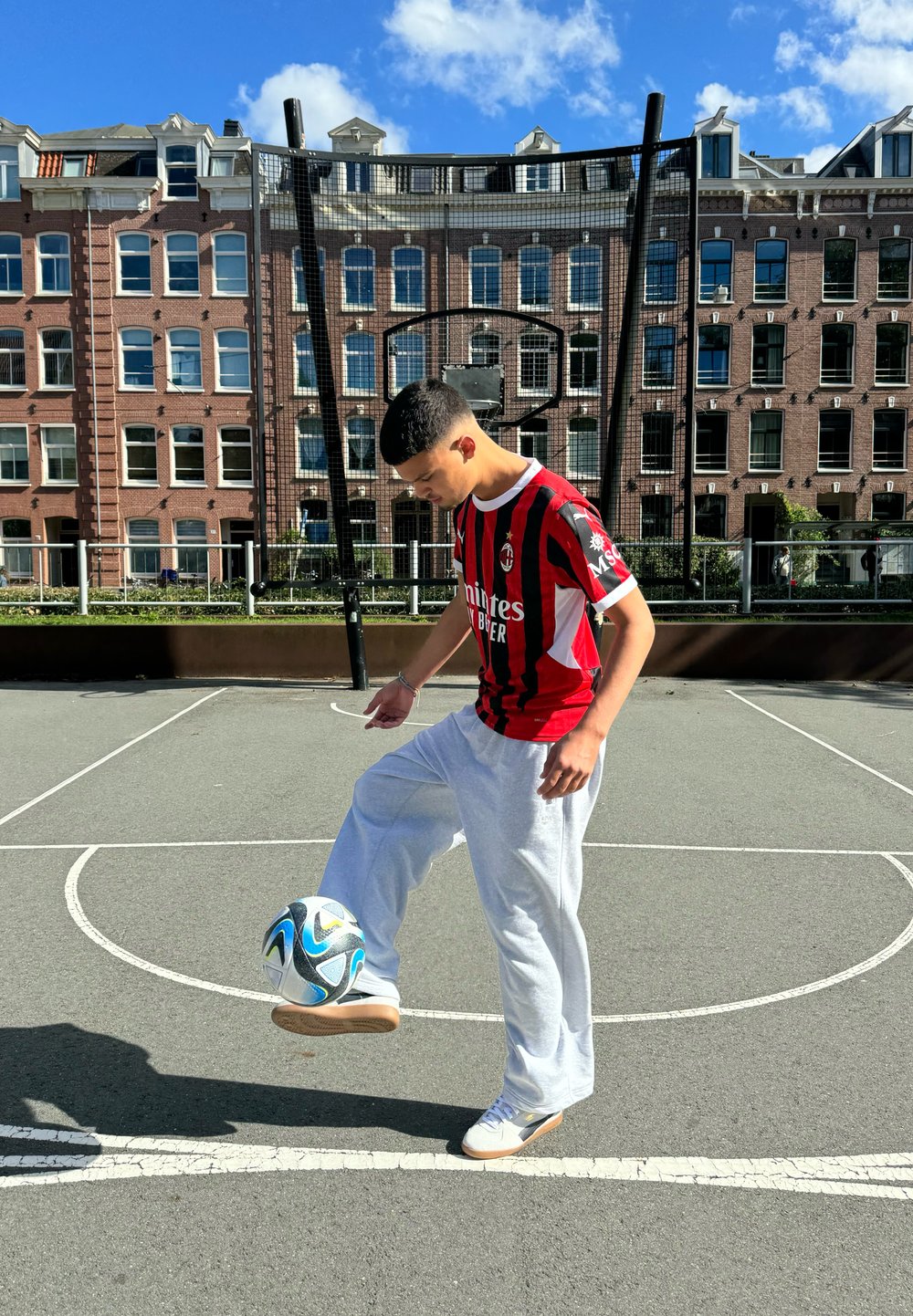 Pessoa vestindo uma camisola listrada horizontalmente em vermelho e preto e calças de moletom cinza claro equilibra uma bola de futebol no pé, em um campo asfaltado.