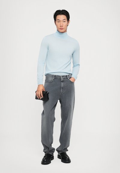 Suéter de cuello alto azul claro, jeans de mezclilla gris, zapatos negros, sosteniendo un bolso negro pequeño. Textura suave, corte casual y diseño minimalista.