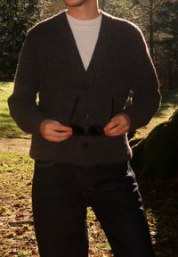 Personne en plein air tenant des lunettes de soleil noires, portant un cardigan sombre par-dessus une chemise blanche et un pantalon foncé, avec la lumière du soleil filtrant à travers les arbres.