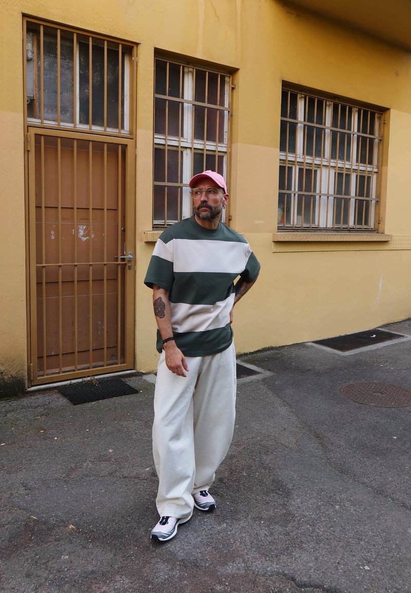 Homme portant une casquette rose, une chemise rayée verte et blanche, un pantalon blanc et des baskets, debout dans la rue près d’un bâtiment jaune avec des fenêtres et une porte grillagées.