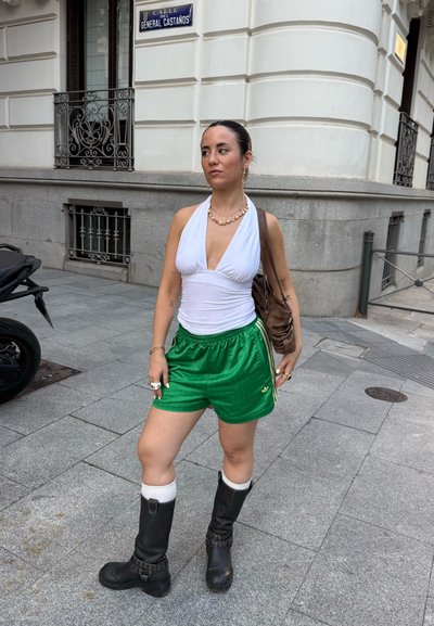 Top halter blanco, shorts deportivos verdes con rayas, botas negras hasta la rodilla, bolso marrón de hombro y accesorios que incluyen anillos y pendientes.