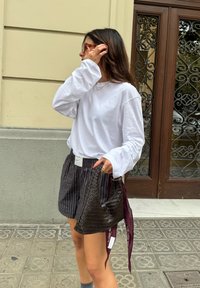 Bolsa tecida preta com um destaque em bordô, usada com uma camisa branca de manga longa e calções às riscas. O fundo apresenta uma porta de madeira ornamentada.