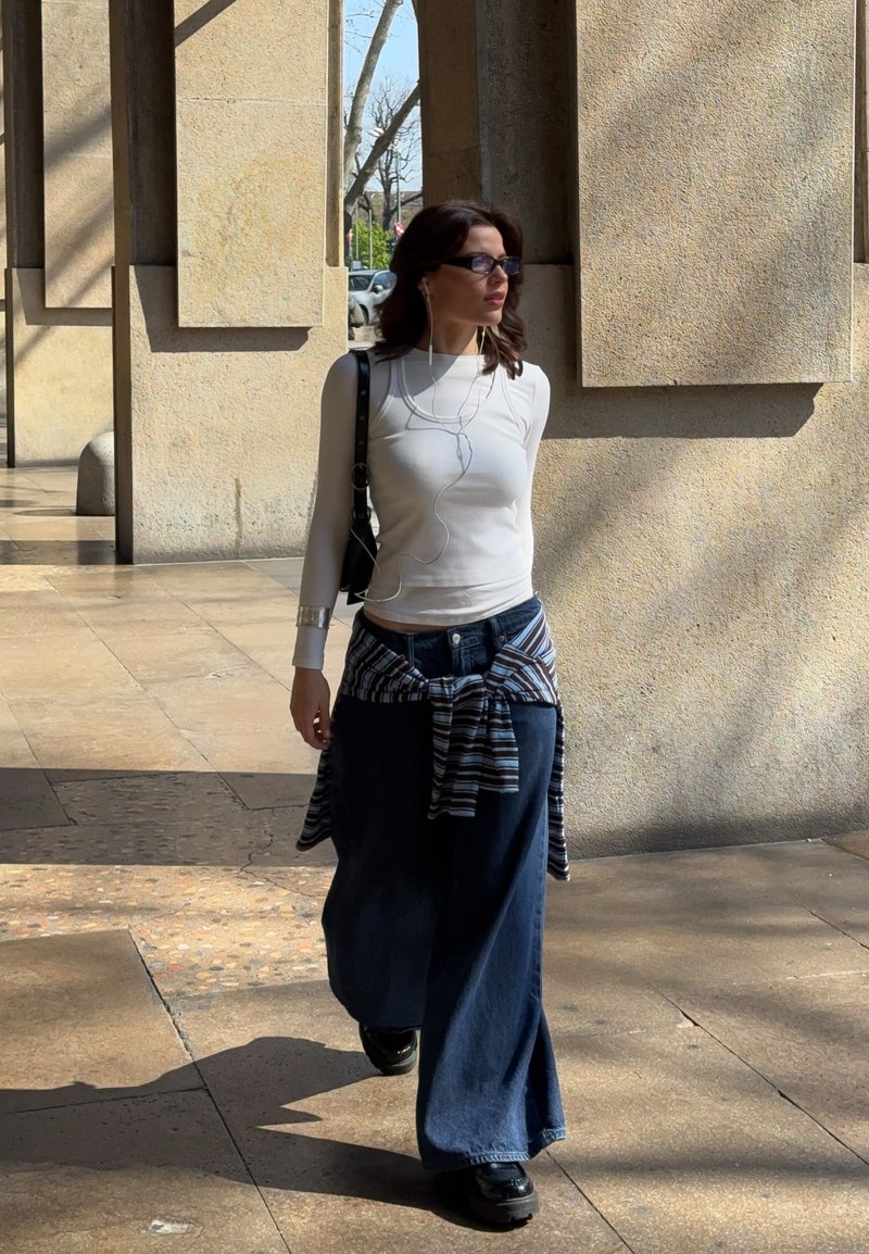 Frau trägt weißes Langarmshirt und weite blaue Jeans, läuft unter hohen Steinsäulen, trägt Sonnenbrille und hört Kopfhörer.