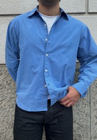 Camicia azzurro chiaro in cotone con bottoni, colletto appuntito e maniche lunghe, indossata sopra una maglietta bianca. Jeans scuri e sfondo con parete testurizzata.