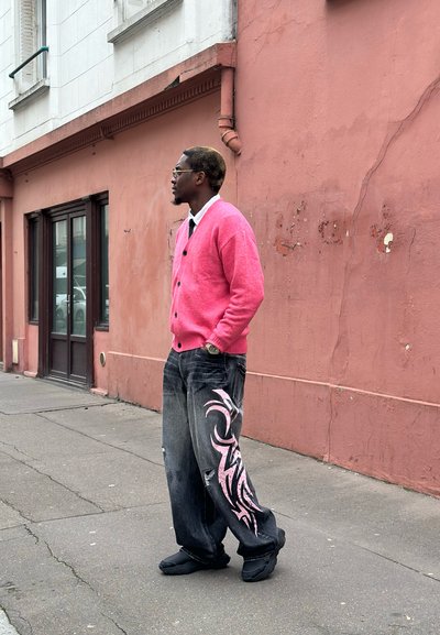 Cardigan rosa claro, camisa blanca con corbata negra, jeans negros desgastados con diseño tribal rosa y calzado negro. Entorno urbano con una pared rosa.