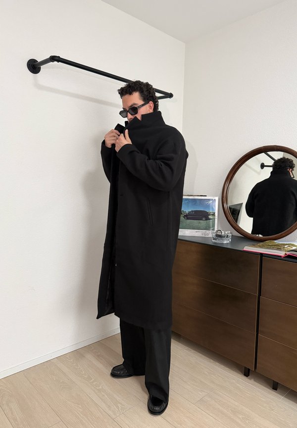 Homme portant des lunettes de soleil noires et un long manteau noir, debout à côté d'une commode en bois avec un miroir rond et un cadre photo dessus.