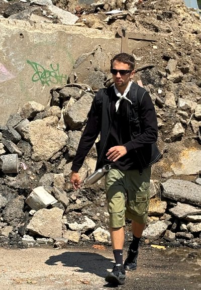 Una figura masculina camina entre escombros de construcción, vistiendo una chaqueta negra, pantalones cortos de carga oliva y zapatos negros, con una botella de agua a su lado.