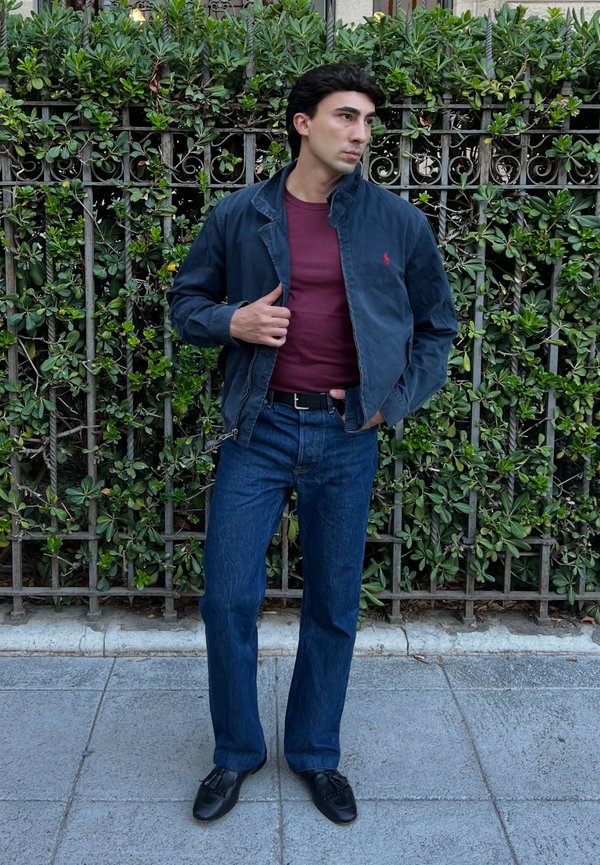 Hombre con chaqueta azul marino, camisa granate, vaqueros azules y zapatos negros, de pie sobre la acera frente a una valla verde y frondosa.