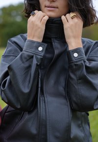 Jachetă din piele neagră cu guler înalt și manșete cu nasturi. Are o textură netedă și o croială ușor supradimensionată.