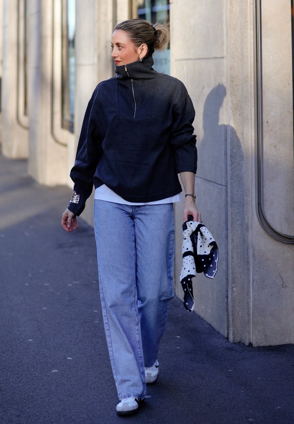 Femme marchant sur le trottoir portant un pull sombre, un jean bleu clair, des baskets blanches, tenant une écharpe à pois noirs et blancs dans sa main droite.