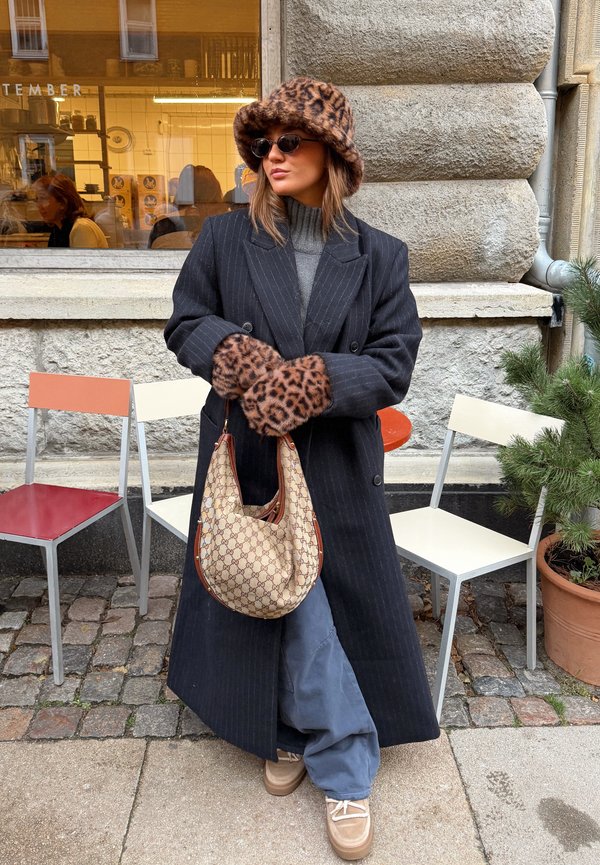 Nainen, joka pukeutunut leopardikuvioiseen hattuunsa ja hansikkaisiin, mustaan viivakuosiin takkiin, harmaisiin housuihin, aurinkolaseihin ja pitää kädessään beigeä kuviollista käsilaukkua kaupungin jalkakäytävällä.