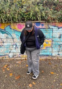 Oseba, oblečena v mornarsko modro kapuco preko črne majice, s sivimi kratkimi hlačami in črno-belimi supergami, stoji pred barvito steno z grafiti.