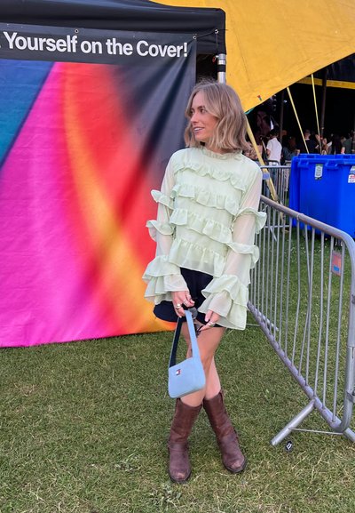 Blusa verde claro con volantes y tela transparente, mangas largas con volantes, combinada con pantalones cortos oscuros y botas de cuero marrón, sosteniendo un pequeño bolso azul.