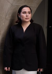 Czarna marynarka z głębokim dekoltem w literę V, wyraźnie zarysowanymi ramionami i dwoma przednimi kieszeniami, w połączeniu z luźną białą odzieżą. Gładka tekstura materiału.