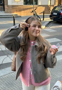 Jonge vrouw met koptelefoon die roze blush aanbrengt met een make-upkwast terwijl ze op een stoep in de stad staat, nabij een zebrapad.
