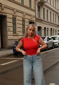 Rode aansluitende t-shirt, lichtblauwe hoge taille jeans en gouden sieraden. De achtergrond toont een stedelijke straat met geparkeerde auto's en gebouwen.