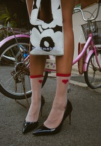 Tacones negros altos sobre piernas que llevan calcetines ligeros y transparentes con dibujos de corazones rojos. Una bolsa de estampado está colgada del brazo. En el fondo hay una bicicleta rosa.