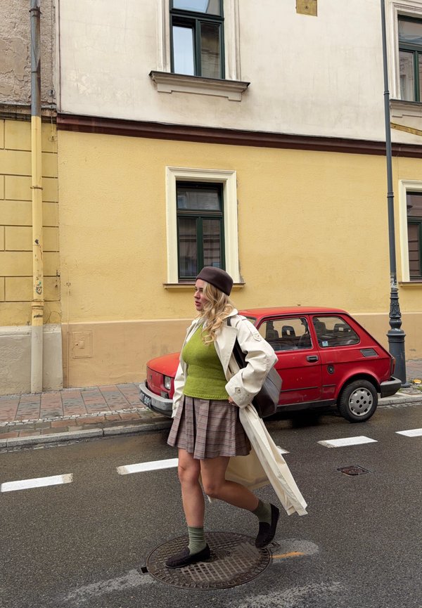 Femme en pull vert, jupe à carreaux, manteau beige et béret marchant dans une rue mouillée près d'une voiture rouge et d'un bâtiment jaune avec des fenêtres.