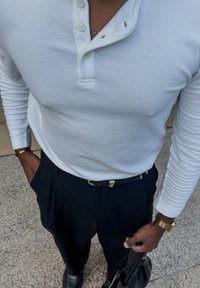Camisa branca de manga longa tipo henley com tecido texturizado, três botões no pescoço; combinada com calças pretas de cintura alta e segurando uma bolsa preta.