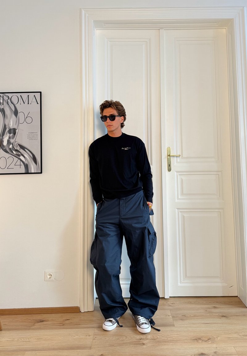 Maglietta nera a maniche lunghe, pantaloni cargo blu navy, sneakers grigie. Occhiali da sole, in piedi contro una porta a pannello bianca con un poster incorniciato nelle vicinanze.
