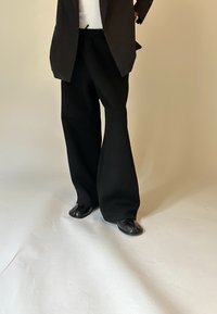 Person in schwarzen Schlaghosen, schwarzer offener Jacke, weißem Oberteil und schwarzen Zehenschuhen, steht auf einer hellen Oberfläche.