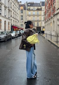 Veste en cuir marron, jean large bleu clair, et sac texturé vert citron avec des accents tressés, debout dans une rue mouillée.