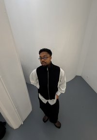 Fekete kötött mellény fehér hosszú ujjú ing felett, laza fekete nadrággal és sötét cipővel párosítva. A háttérben fehér falak és szürke padló látható.