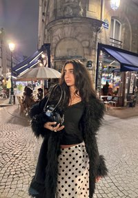 Abrigo de piel sintética negra, top negro y falda blanca con lunares. Sosteniendo un bolso negro, de pie en una calle adoquinada por la noche.