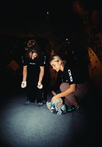 Due scalatori si preparano in una palestra di arrampicata, uno indossa scarpe da arrampicata nere e ha il magnesio sulle mani, l'altro tiene una borsa a motivo leopardato.