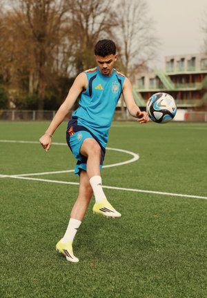 Joven atleta masculino con ropa deportiva azul pateando un balón de fútbol en un campo de césped con árboles y un edificio al fondo.