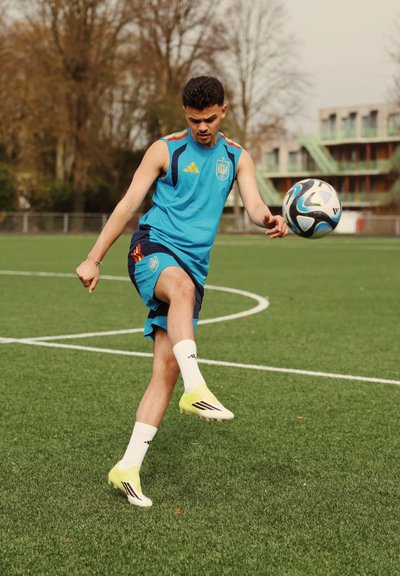 Joven atleta masculino con ropa deportiva azul pateando un balón de fútbol en un campo de césped con árboles y un edificio al fondo.