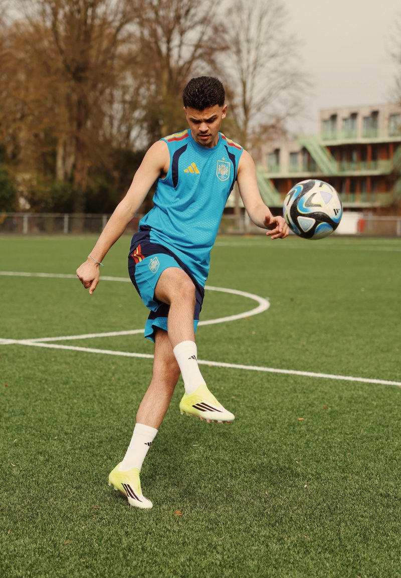 Jeune athlète masculin en tenue de sport bleue donnant un coup de pied à un ballon de football sur un terrain herbeux avec des arbres et un bâtiment en arrière-plan.