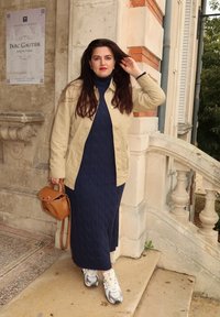 Beige jakke over en mørk blå kabelstrikket kjole, kombinert med hvite sko. Brun håndveske med rødt aksentdetalj. Utendørs omgivelser ved en bygning.