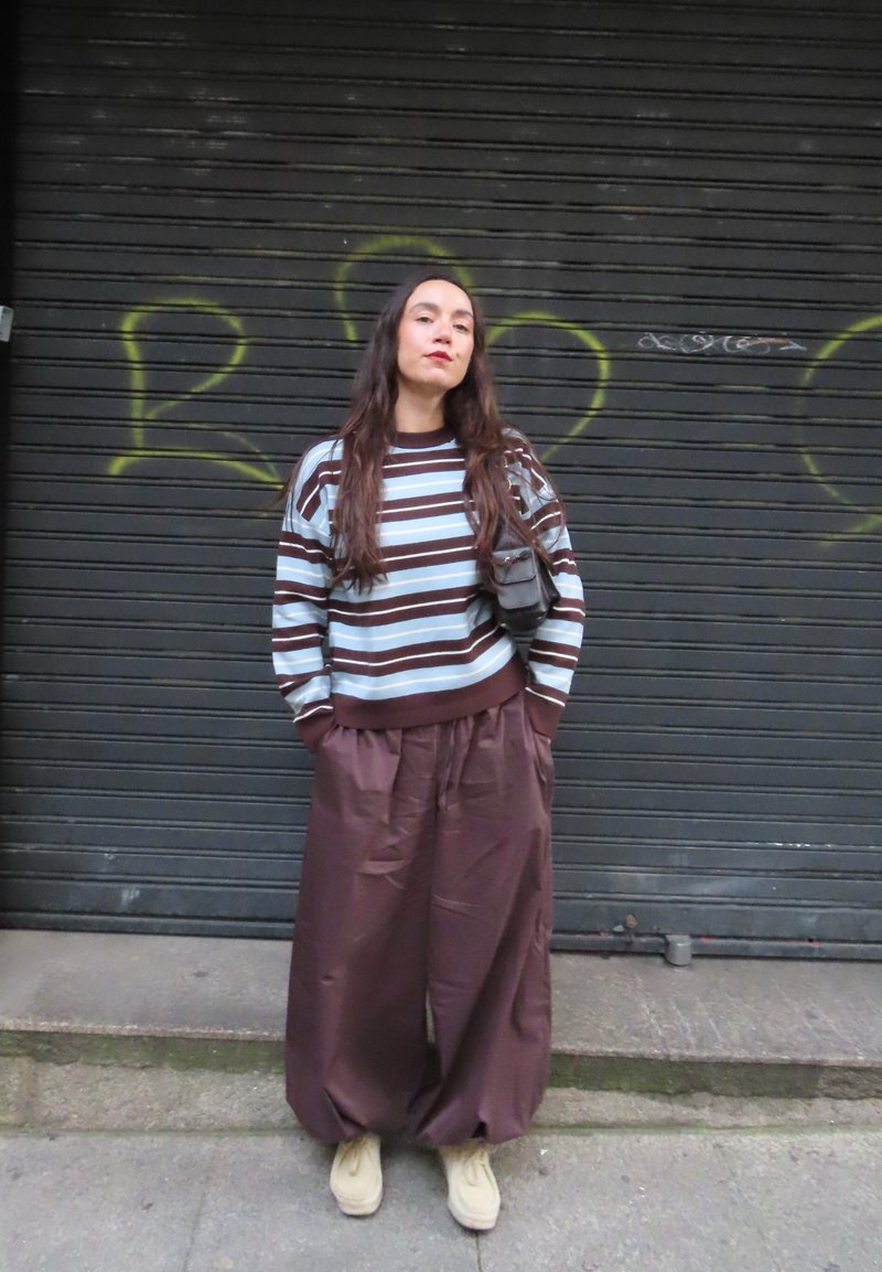 Giovane donna con i capelli lunghi in piedi davanti a una saracinesca nera coperta di graffiti, indossa un maglione a righe, pantaloni marroni larghi e scarpe beige.