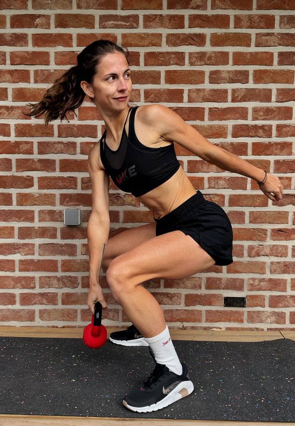 Una mujer con ropa deportiva realiza un balanceo bajo con una pesa rusa en un gimnasio, sosteniendo una pesa rusa roja junto a una pared de ladrillo.