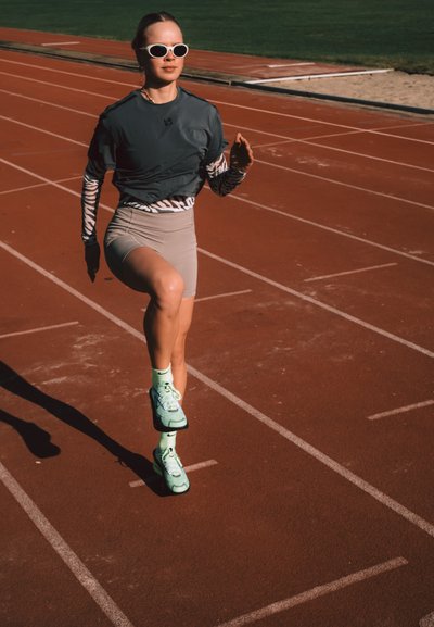 Mujer con gafas de sol y ropa deportiva corriendo en una pista exterior roja con césped al fondo.